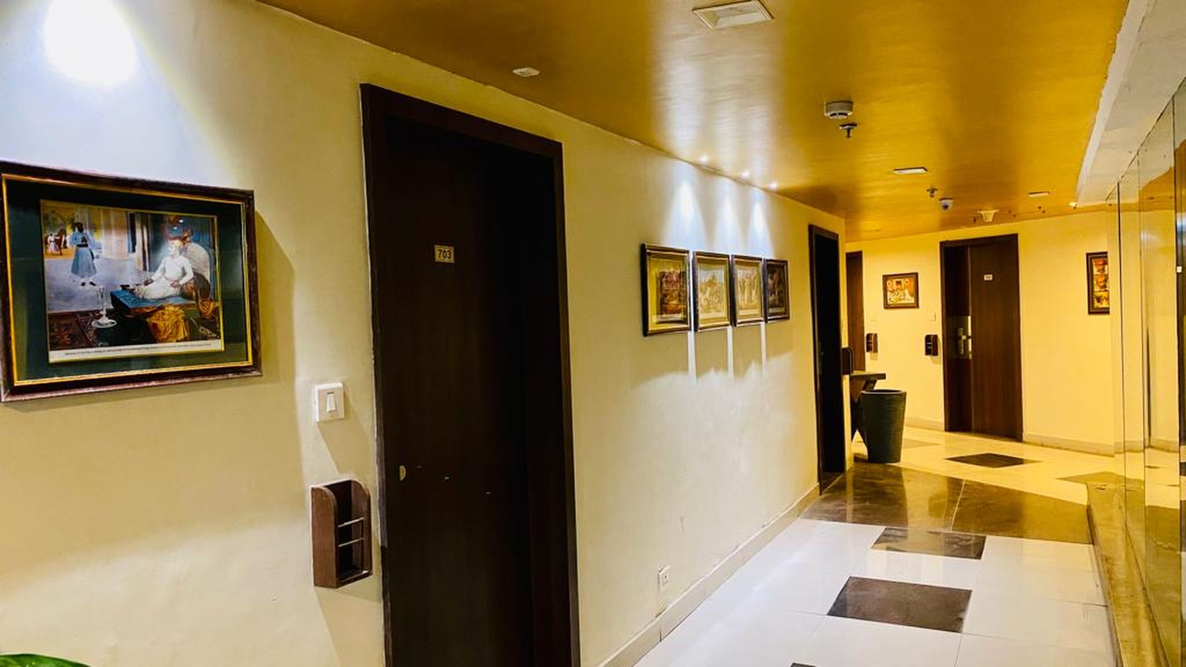 A hotel corridor with a dark door, framed artwork on the walls, and a check-in counter in the distance.