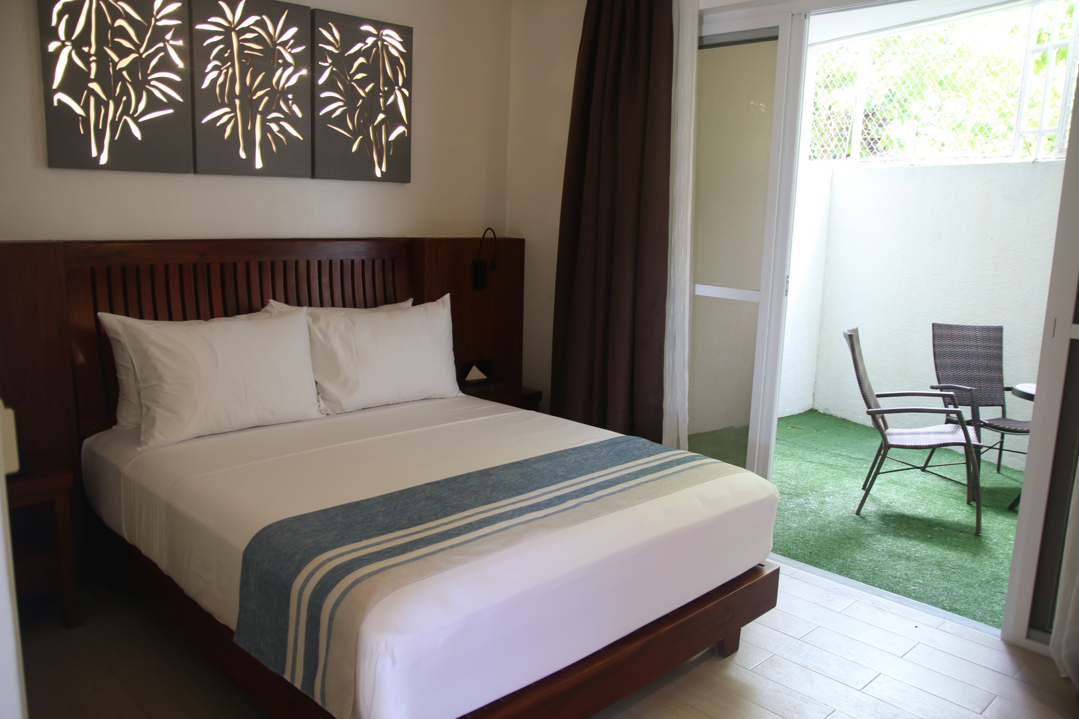 A hotel room with a large bed, a wooden headboard, a wall art piece, and a view of an outdoor area with a chair and a table.