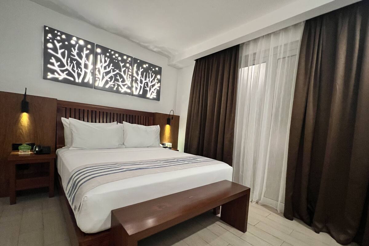 Ein Hotelzimmer mit einem großen Bett, einem Holz-Kopfteil, zwei Nachttischen, einer Bank, einer Wandleuchte und einem Fenster mit Vorhängen.