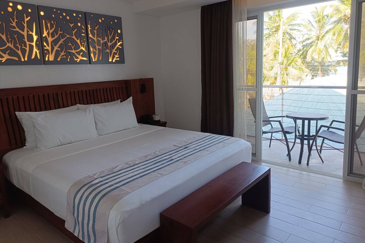 Ein Hotelzimmer mit einem großen Bett, einem hölzernen Kopfteil, einer Bank, einem Nachttisch, einem Wandbild und einem Balkon mit Tisch und Stühlen.
