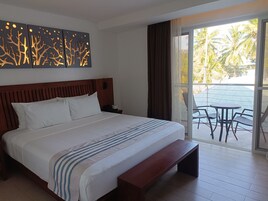Una habitación de hotel con una cama grande, cabecera de madera, un banco, una mesita de noche, una obra de arte en la pared y un balcón con mesa y sillas.