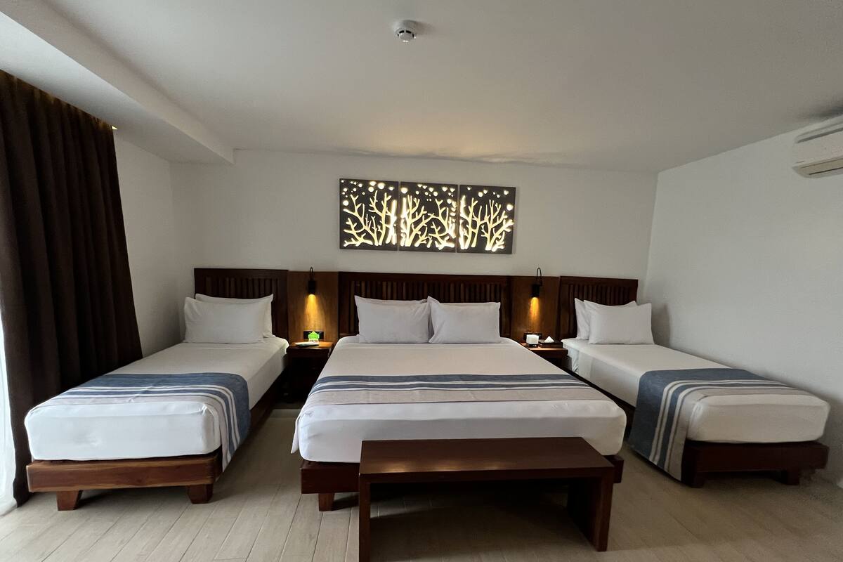 Ein Hotelzimmer mit drei Einzelbetten, einer hölzernen Bank, einem Wandbild und einer Deckenleuchte.