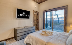 Un dormitorio con una cama, un aparador, un televisor montado en la pared y un ventanal con vistas a árboles.