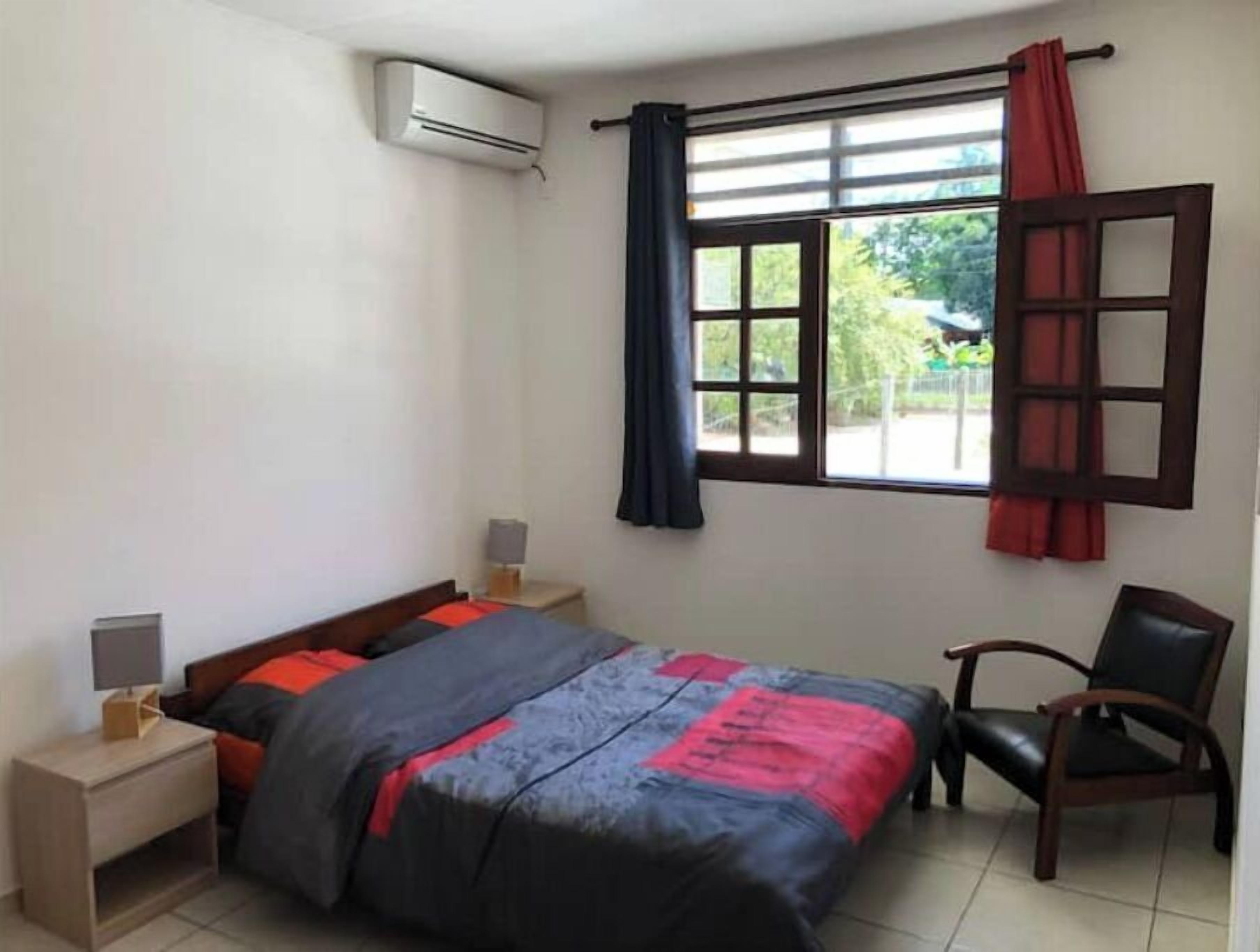 Un dormitorio con una cama, una silla, una mesita de noche, un aire acondicionado y una ventana con cortinas.