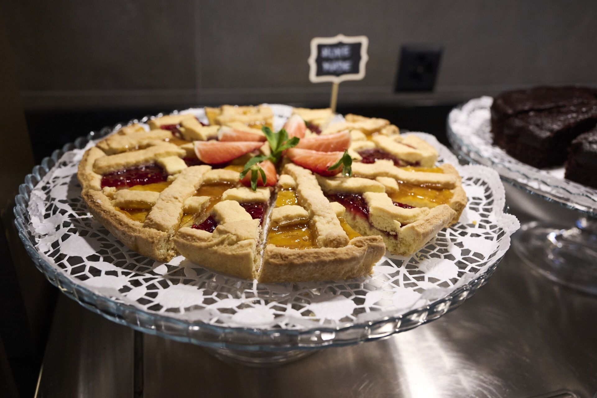 A pie with a lattice crust on a glass plate with a doily, placed on a metal counter.