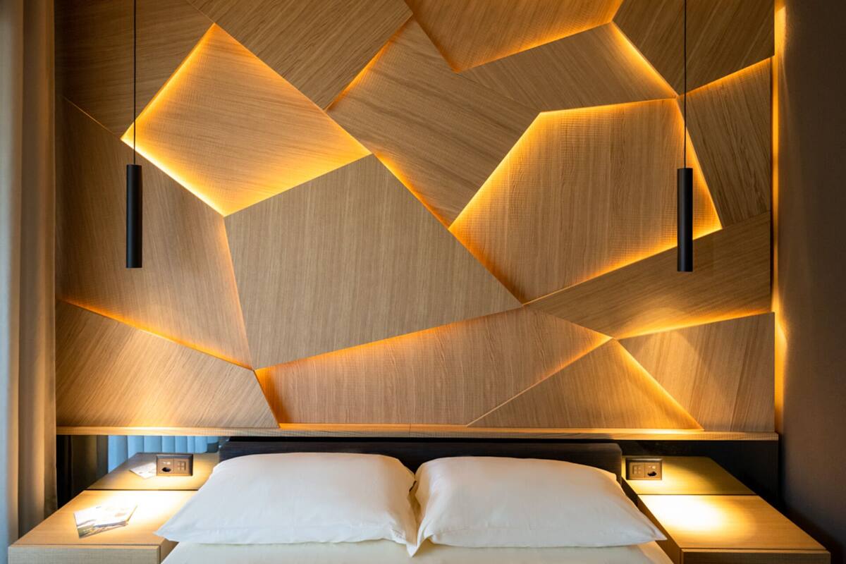 Une chambre d’hôtel moderne avec un mur en bois au motif géométrique, un lit recouvert de linge blanc et deux tables de chevet avec des lampes.
