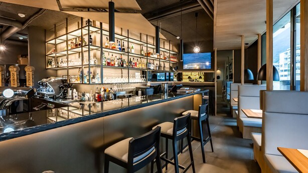 A modern bar area with a long counter, multiple shelves of bottles, and bar stools.