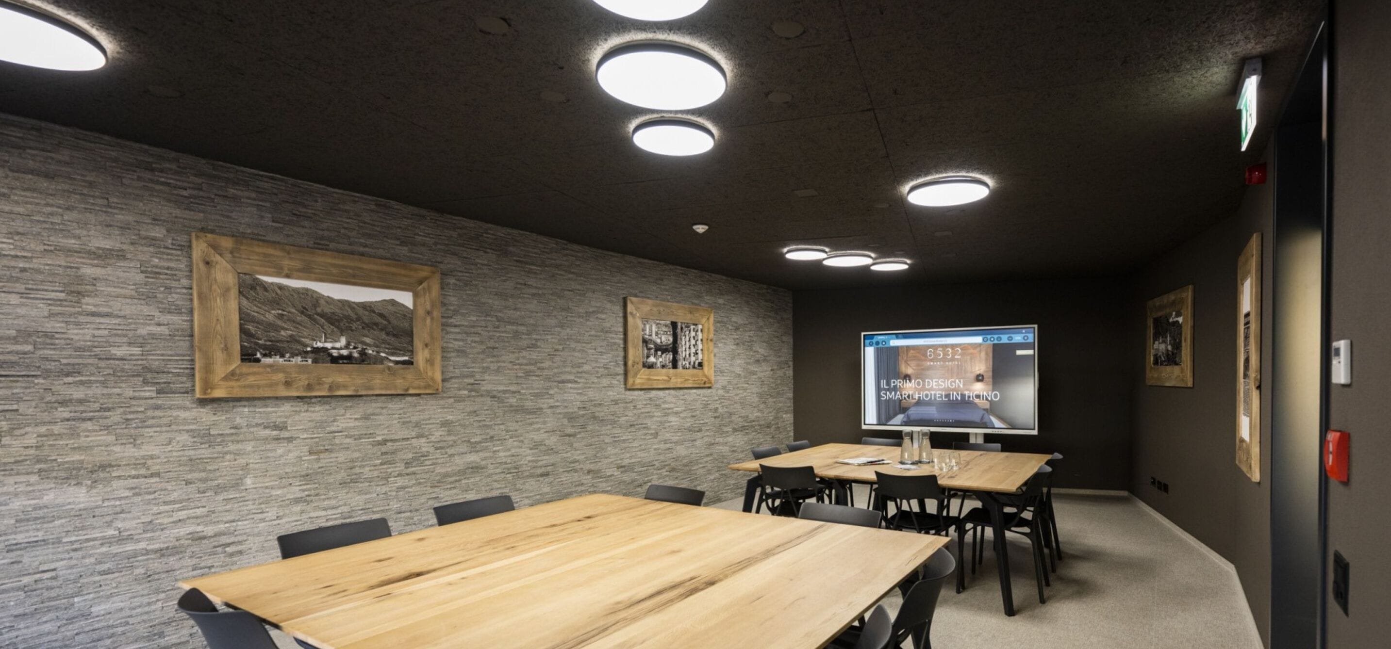 Ett konferensrum med bord och stolar i trä, inramade tavlor på väggen och en stor skärm som visar text.