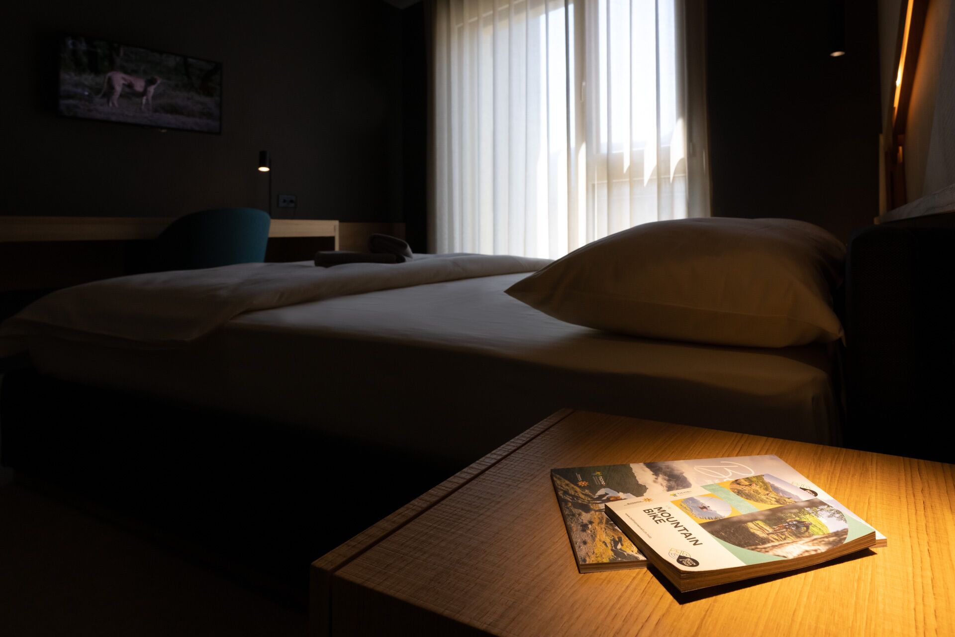 Ein Hotelzimmer mit einem Bett, einem Nachttisch mit einem Buch, einer Lampe und einem Fenster mit Vorhängen.