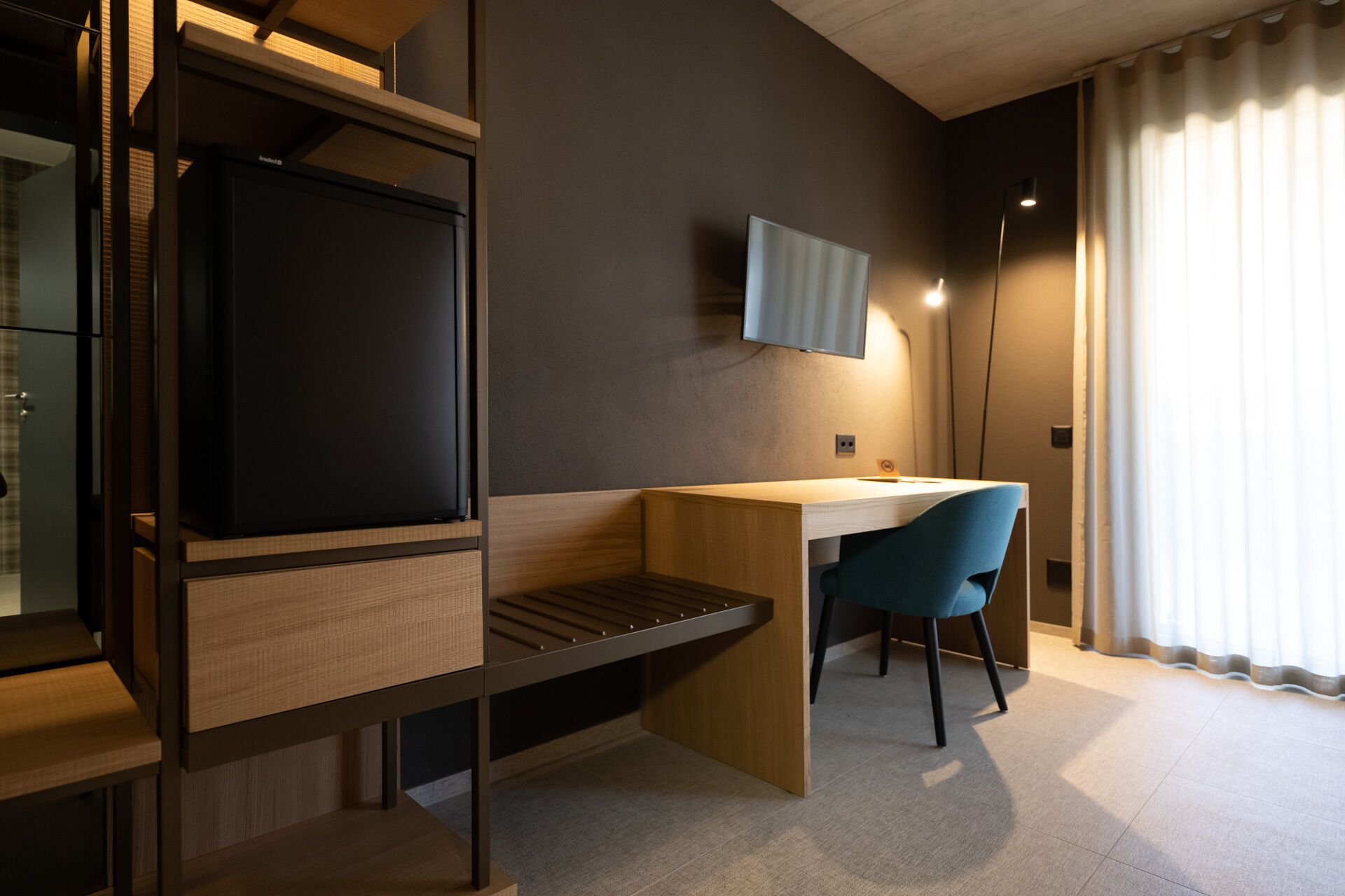 A modern room with a flat-screen TV mounted on the wall, a wooden desk, a blue chair, and a large window with curtains.