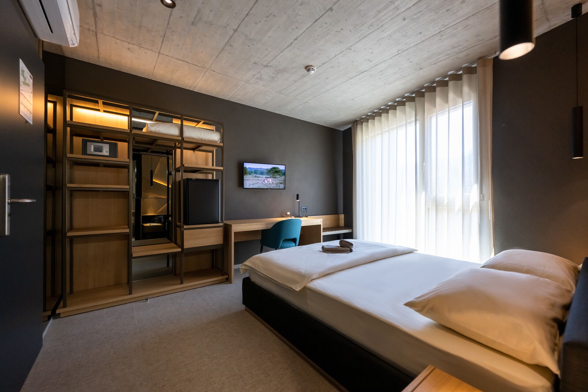 A modern hotel room with a bed, a desk, a chair, and a wardrobe.