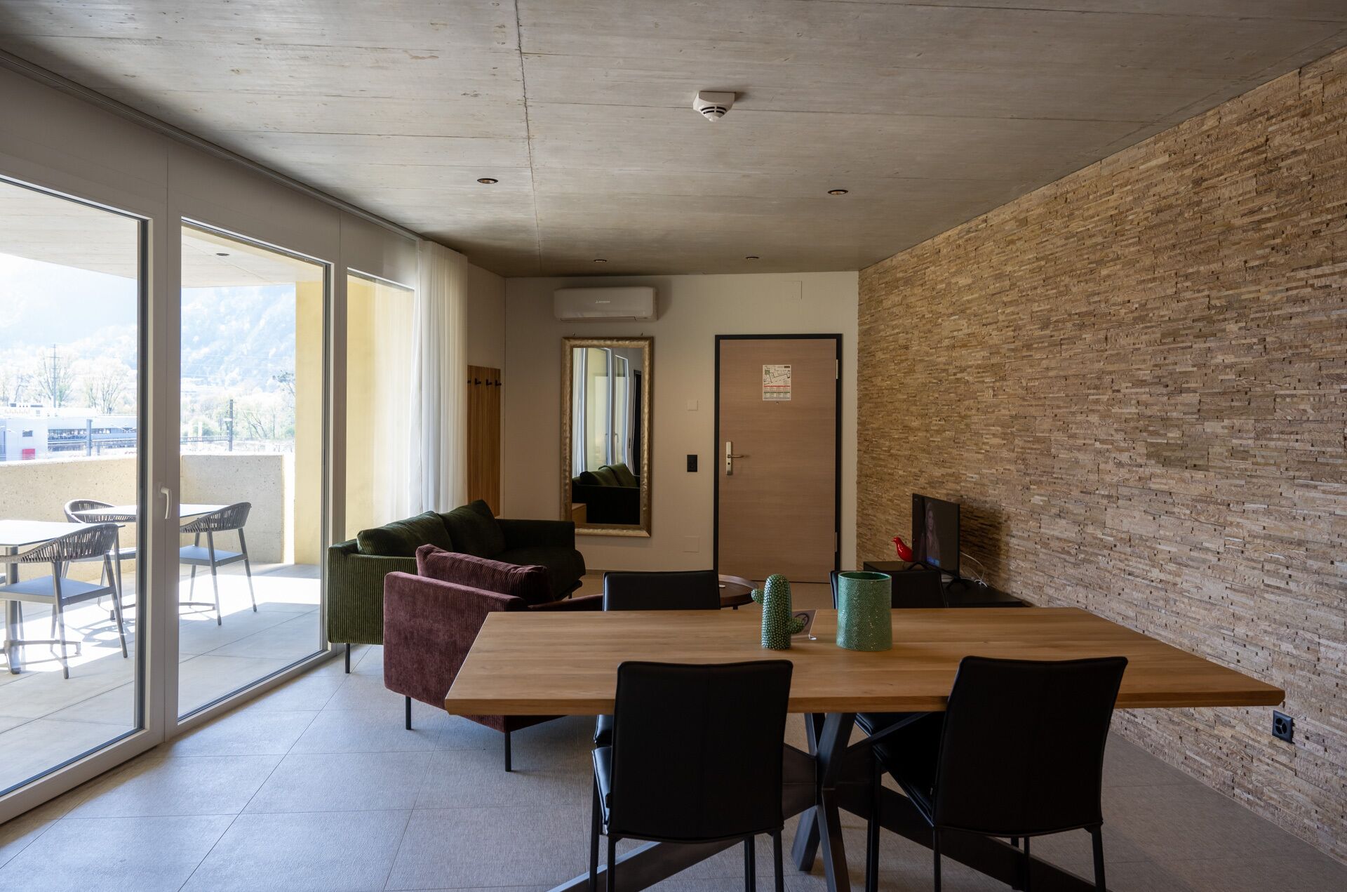A modern interior with a wooden dining table, black chairs, a green sofa, and a large window offering a view of the outdoors.