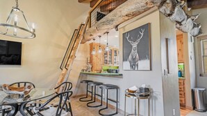 A rustic interior with a dining area, bar stools, a chandelier, and a deer antler decoration.