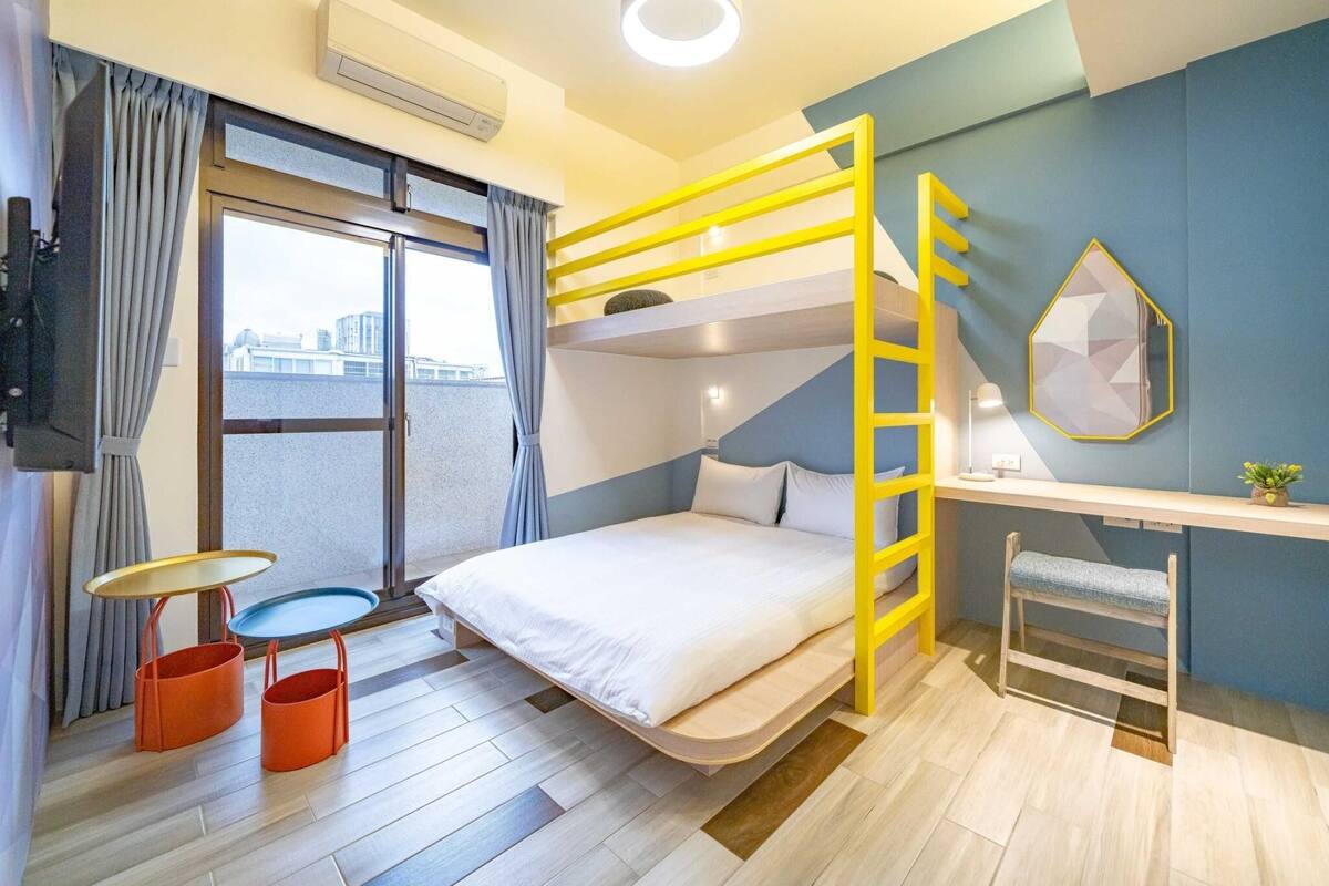 A bunk bed with a yellow ladder, a desk with a lamp, and a view of buildings through a window.