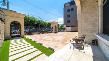 A courtyard with a paved walkway, a small seating area with chairs, and a gate leading to a parking area.