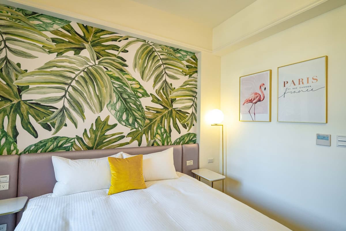 A hotel room with a bed, a nightstand, a wall-mounted lamp, and a tropical leaf-patterned wall. There are two framed pictures on the wall.