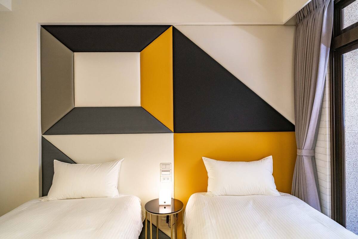 A hotel room with two beds, a geometric wall design, and a window with curtains.