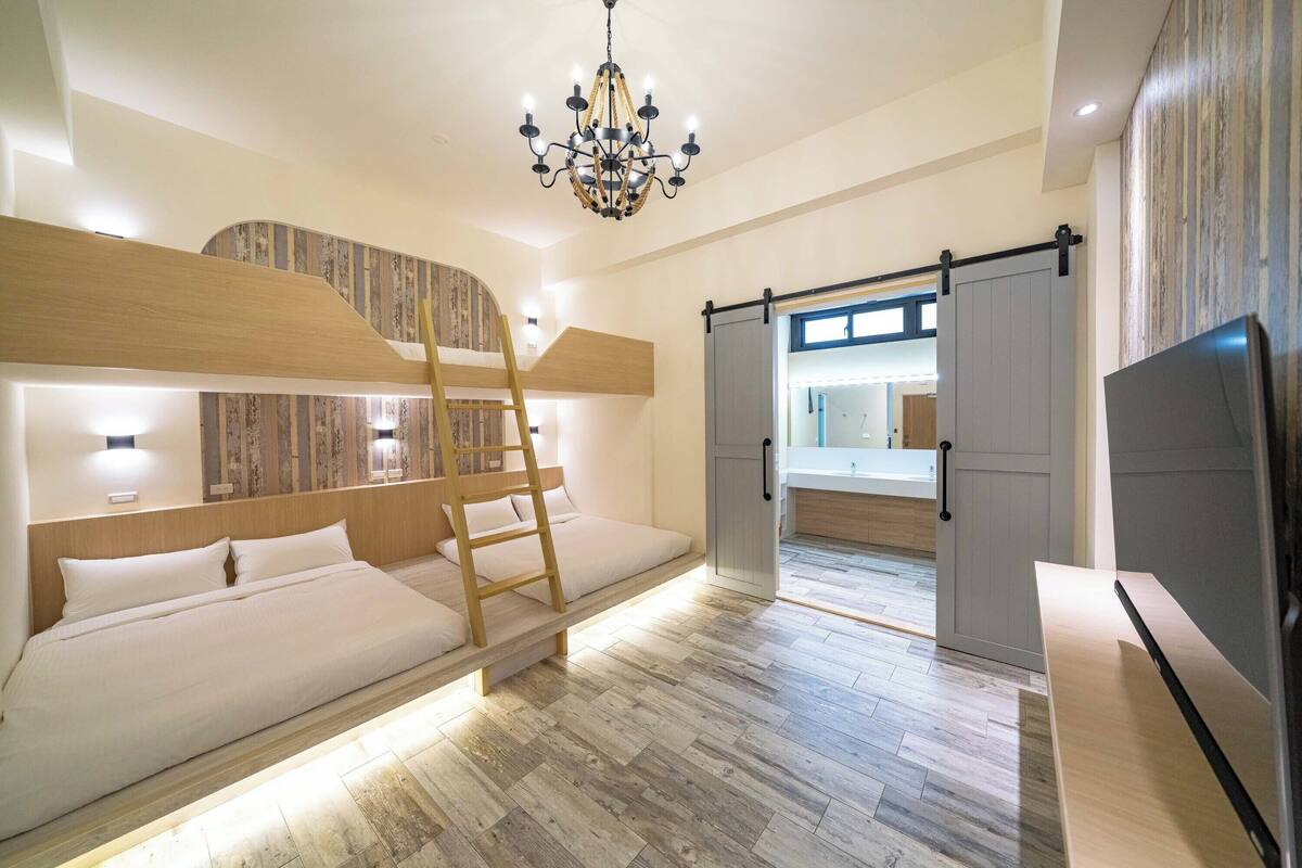 A modern hotel room with bunk beds, a ladder, a chandelier, and a sliding door leading to a bathroom.