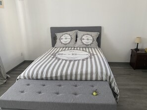 Un lit bien fait, avec une literie à rayures et deux oreillers, un banc recouvert d’un tissu gris et une petite table de chevet avec une lampe.