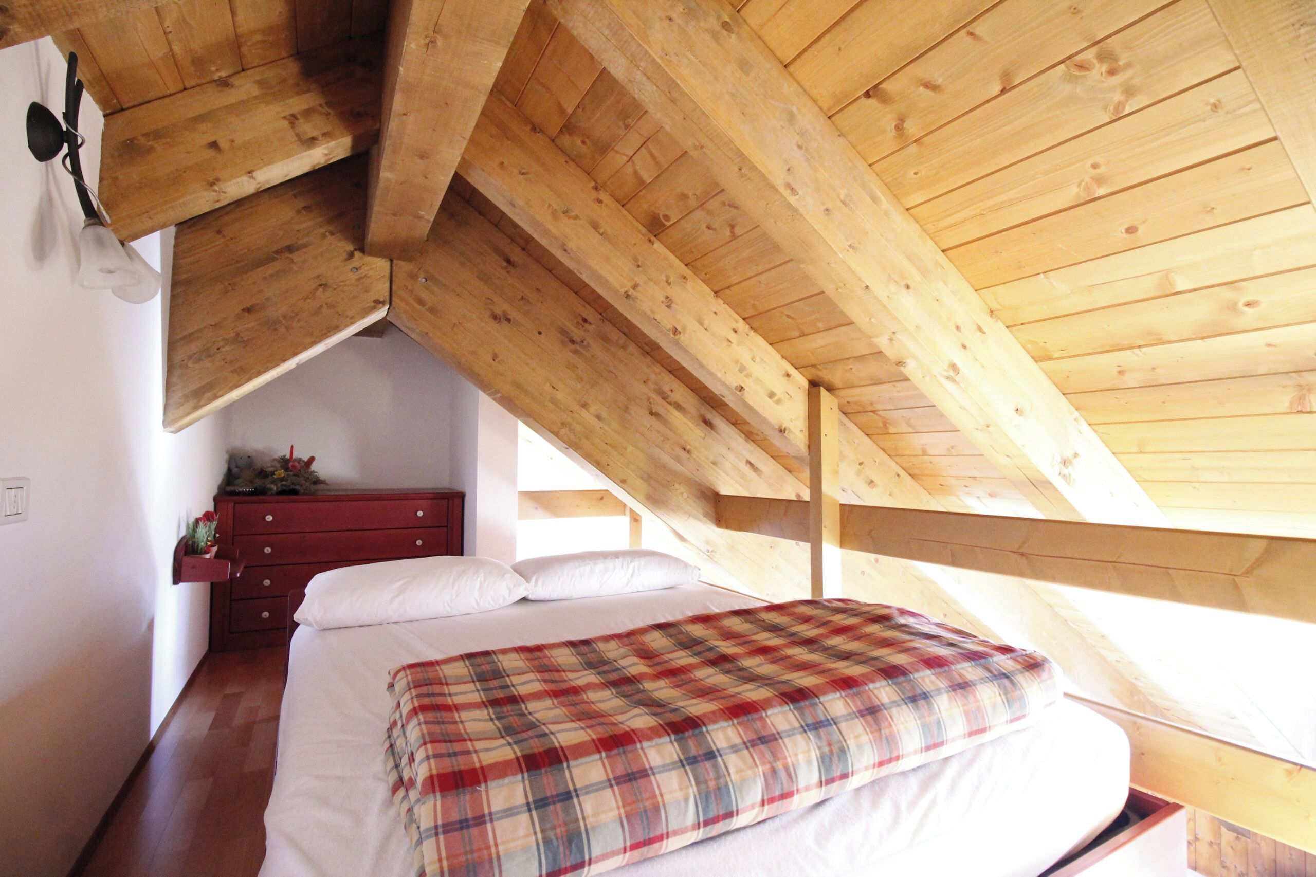 Une chambre avec un plafond en bois, un lit recouvert d’une couverture à carreaux, une commode et une petite plante.