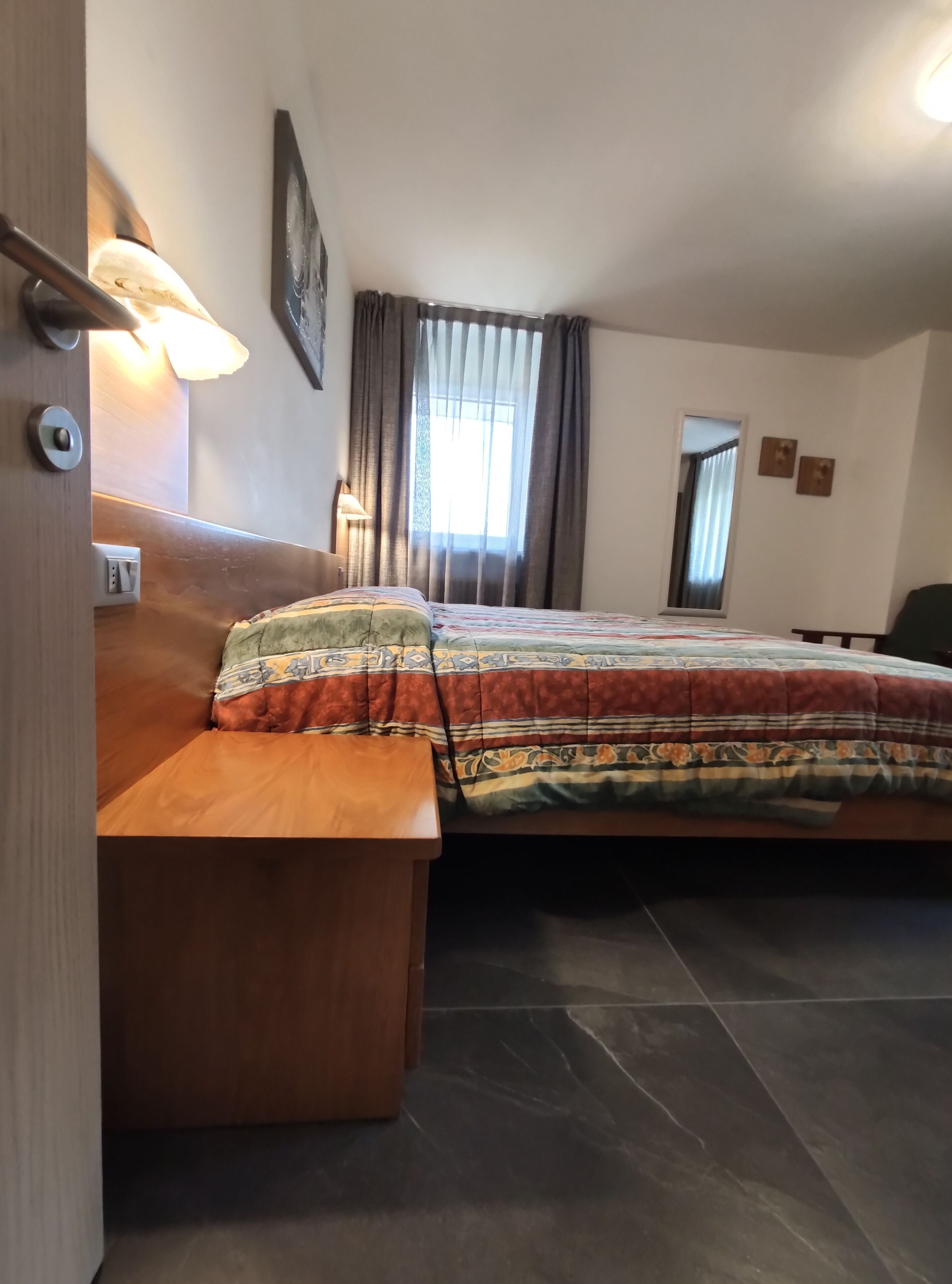 A hotel room with a bed, a wooden nightstand, a lamp, a window with curtains, and a door.