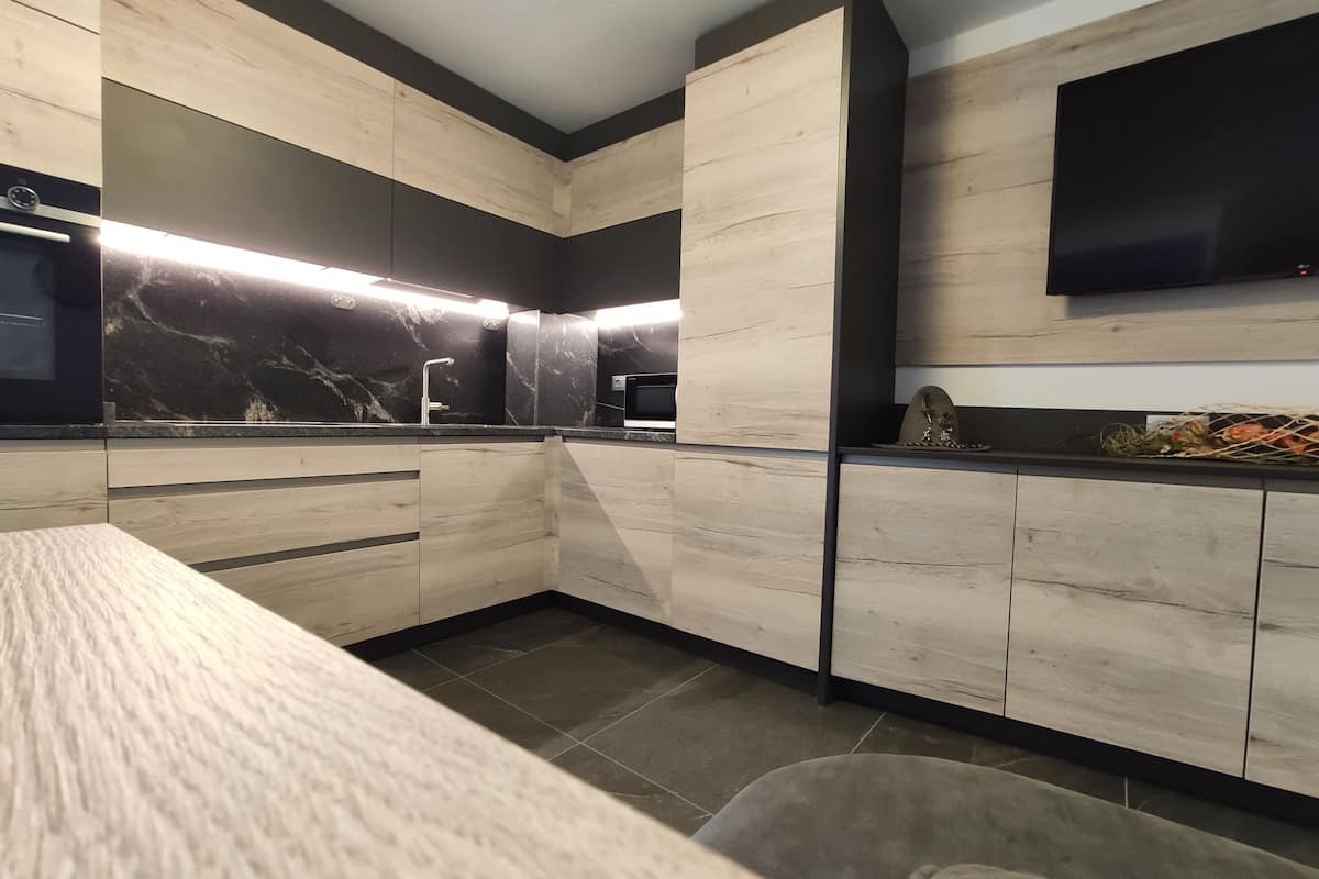 A modern kitchen with wooden cabinetry, a marble backsplash, and a flat-screen TV mounted above the counter.