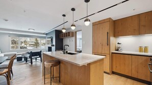 A modern kitchen with wooden cabinets, a central island with stools, pendant lighting, and a view of a snowy landscape through large windows. - Lodge Tower 486 2 BR at the base of Vail Mountain (Vail)