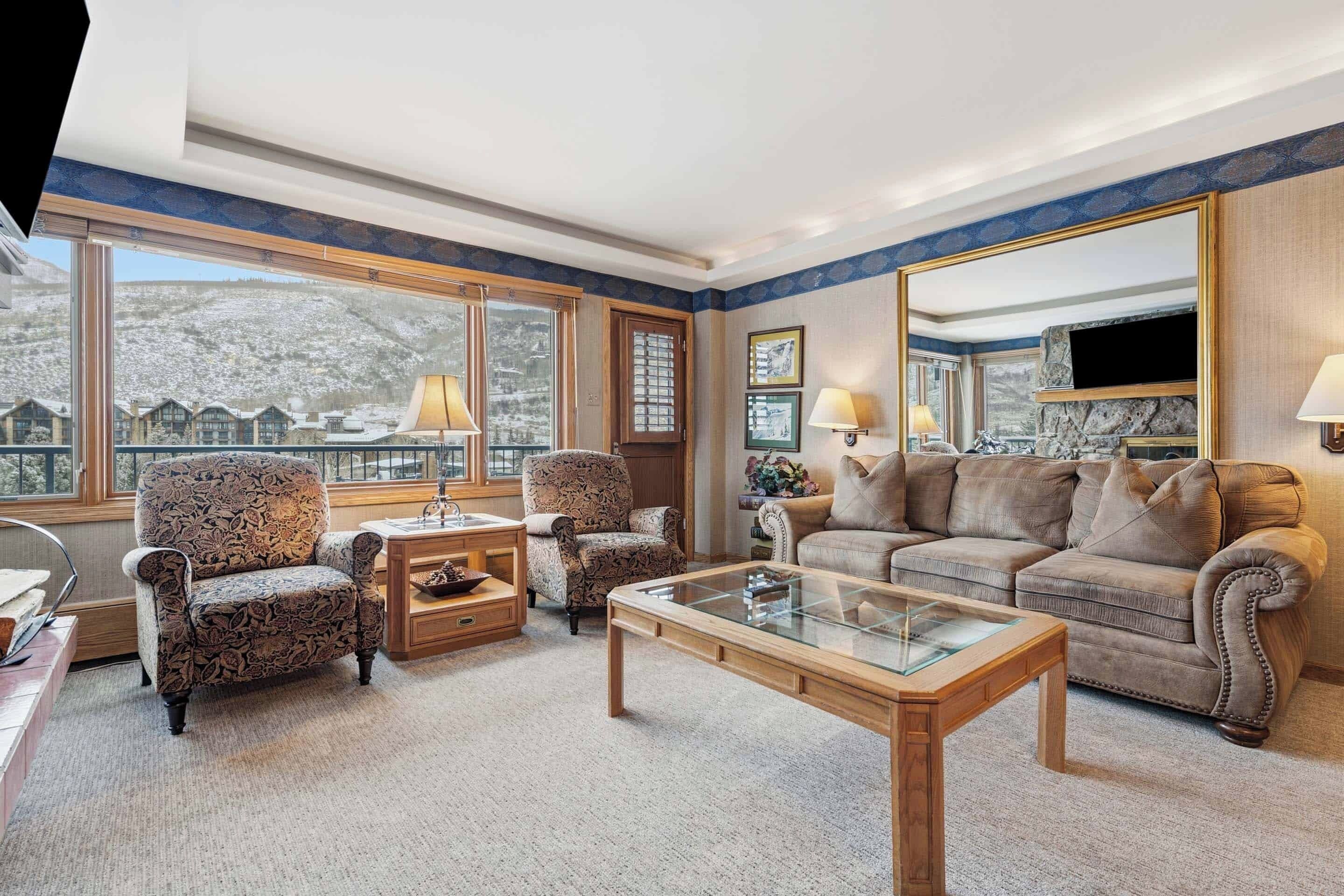 A spacious living room with a large window offering a mountain view, a comfortable sofa, two patterned armchairs, and a glass coffee table.
