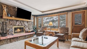 A cozy living room with a stone fireplace, comfortable seating, and a large window offering a snowy mountain view.