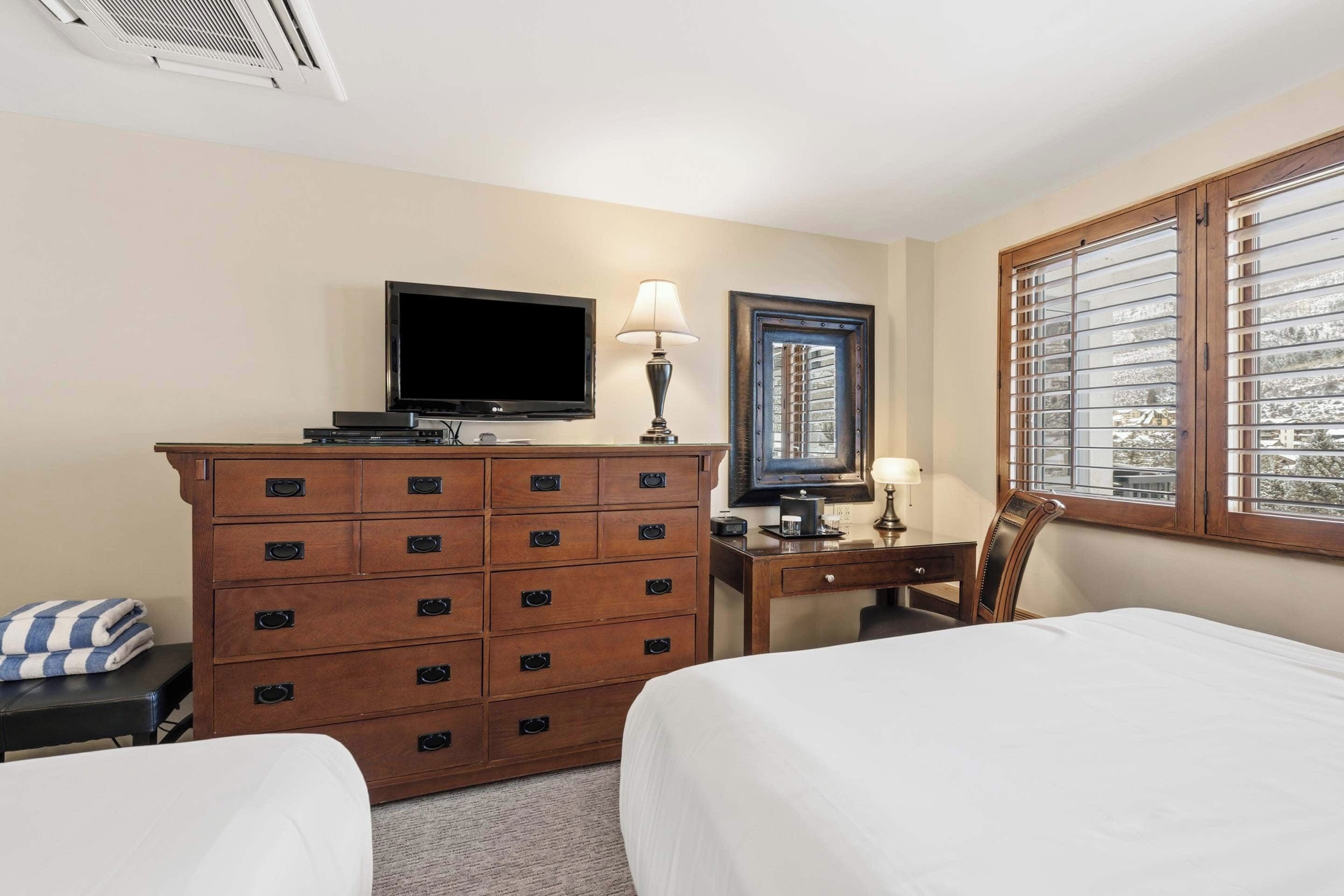 A hotel room with two beds, a TV, a dresser, a desk, and a window with blinds.