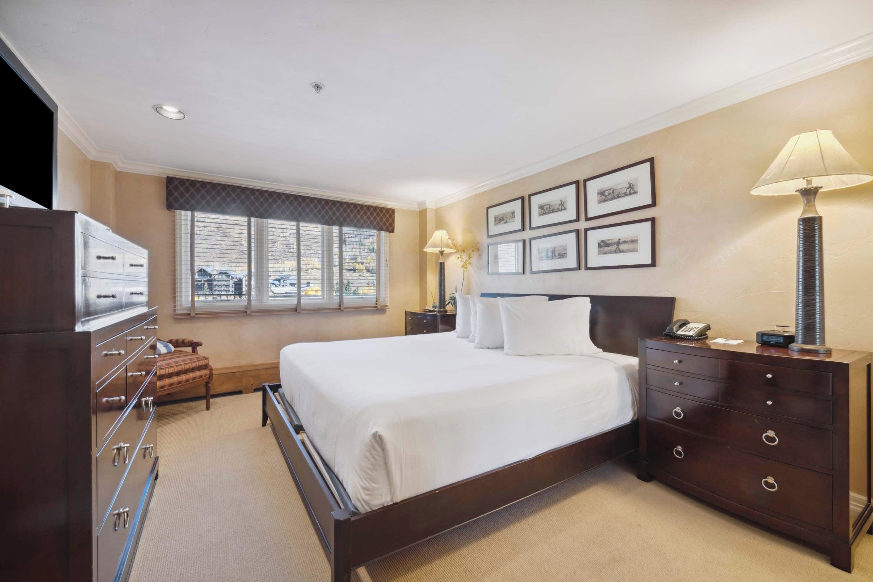 A hotel room with a large bed, a dresser, a nightstand, and a lamp.