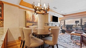 A dining area with a round table and six chairs, a chandelier, a painting on the wall, and a large window with a view.