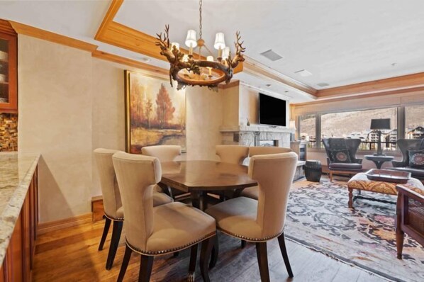A dining area with a round table and six chairs, a chandelier, a painting on the wall, and a large window with a view. - Lodge Tower 694 2BR at the base of Vail Mountain (Vail)