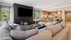 Een moderne woonkamer met een open haard, grijze banken, een flatscreen-televisie en een eetgedeelte met houten kasten en een kroonluchter.