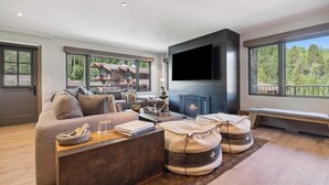 Een moderne woonkamer met een grote bank, een open haard, een aan de muur bevestigde televisie en grote ramen met uitzicht op de omgeving.