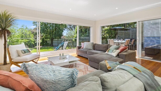Ein geräumiges Wohnzimmer mit einem großen Schiebeglasfenster, das in einen Garten führt, mit einem grauen Sofagarnitur, einem weißen Sessel und einem Couchtisch.