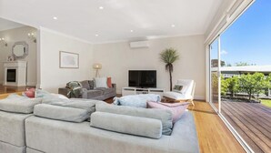 Ein modernes Wohnzimmer mit einem großen grauen Ecksofa, einem Flachbildfernseher und einem Holzdeck, das durch eine große Schiebeglastür sichtbar ist.