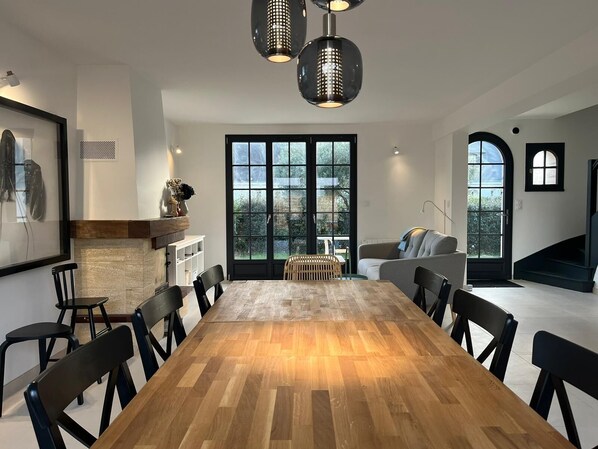 Uma sala de jantar moderna com mesa de madeira e cadeiras pretas, janelas amplas e uma escada que leva a um andar superior.