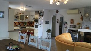 A cozy living room with a wooden dining table and yellow armchair, a kitchen with white appliances and a tiled backsplash, and a ceiling light fixture. - Maison Individuelle de Caractère (Roquebrune-sur-Argens)