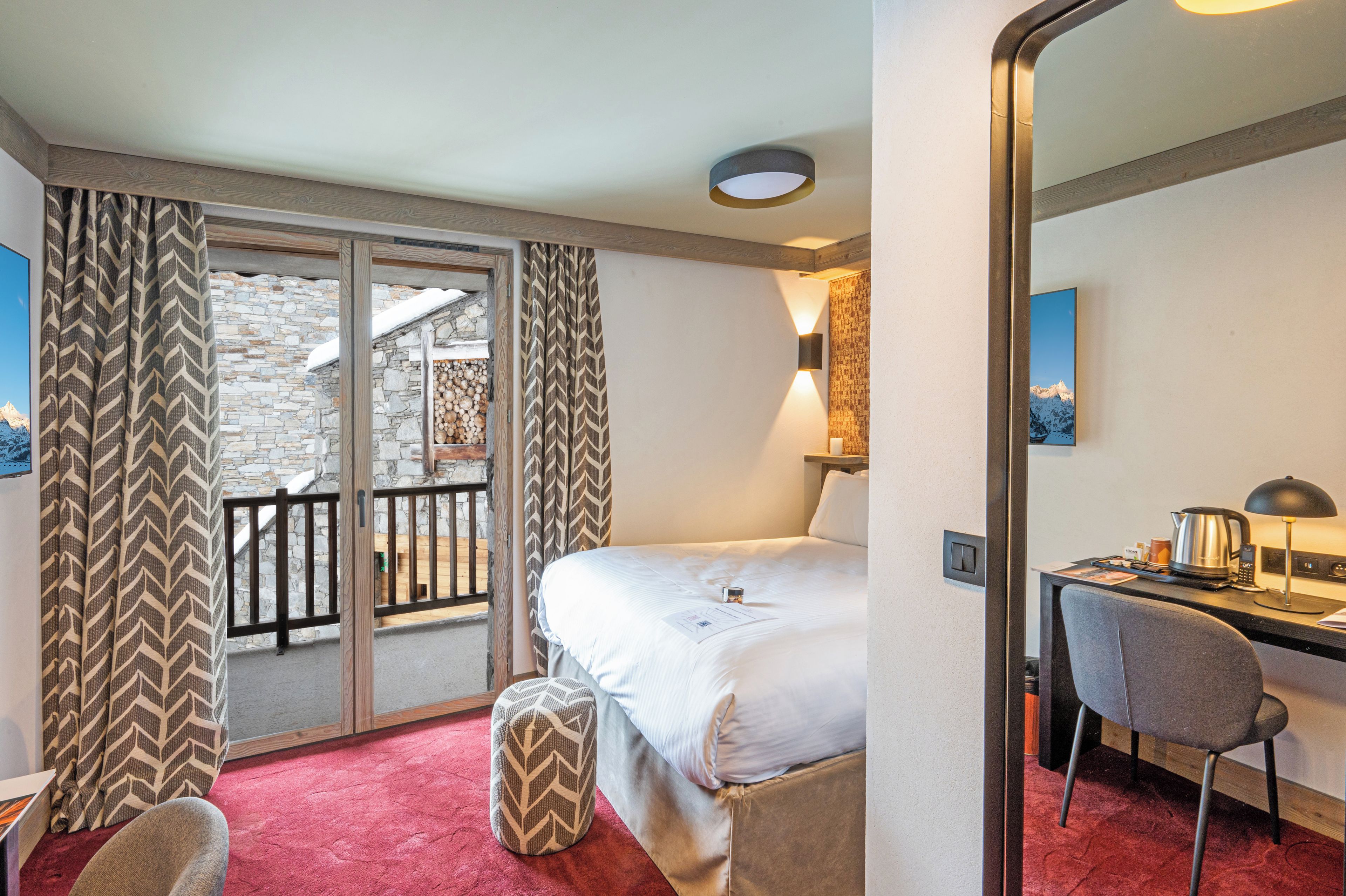 Habitación de hotel con cama, escritorio, silla, alfombra roja y vistas a un muro de piedra y una terraza de madera.