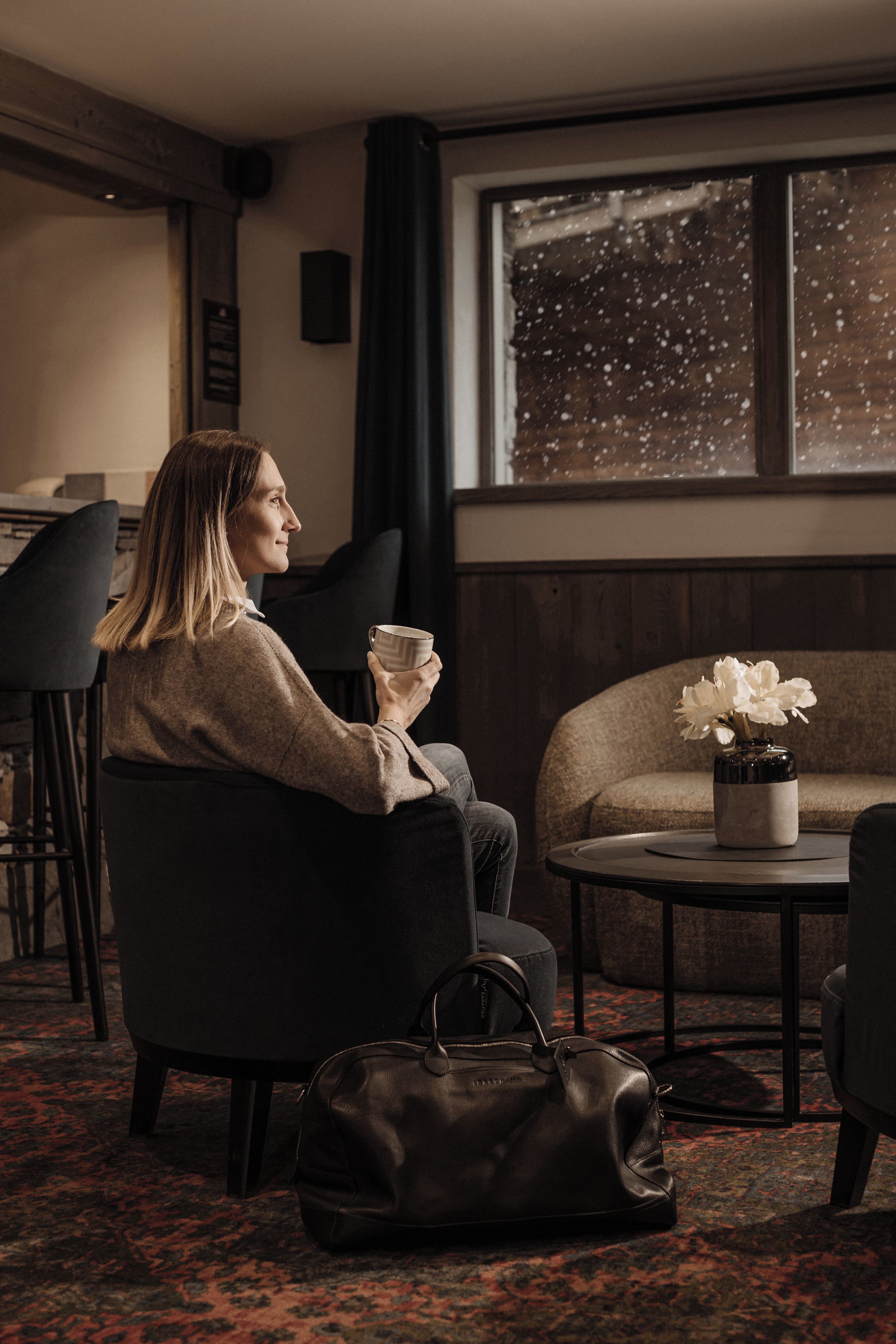 Une personne assise sur une chaise, avec un sac à main posé sur le sol, une table sur laquelle se trouve un vase de fleurs et une fenêtre donnant sur un paysage enneigé.
