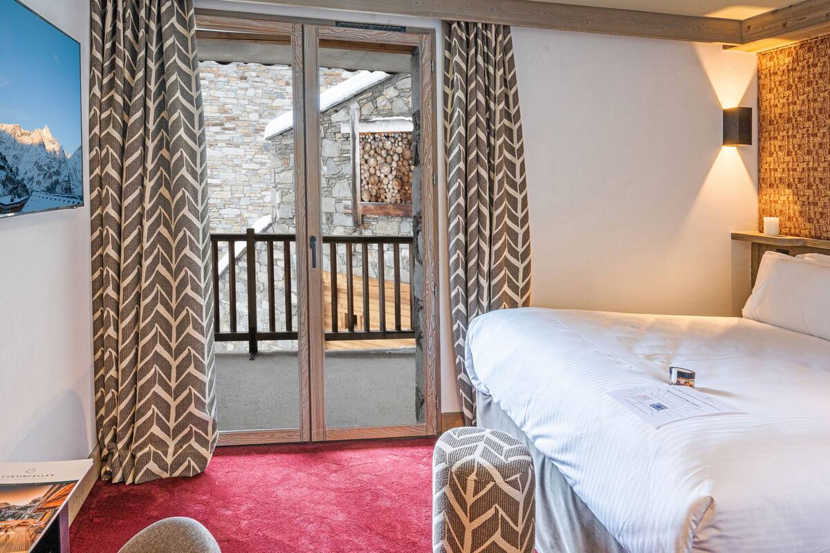 Habitación de hotel con una cama, alfombra roja, ventana con marco de madera y vistas a un muro de piedra y montañas nevadas.