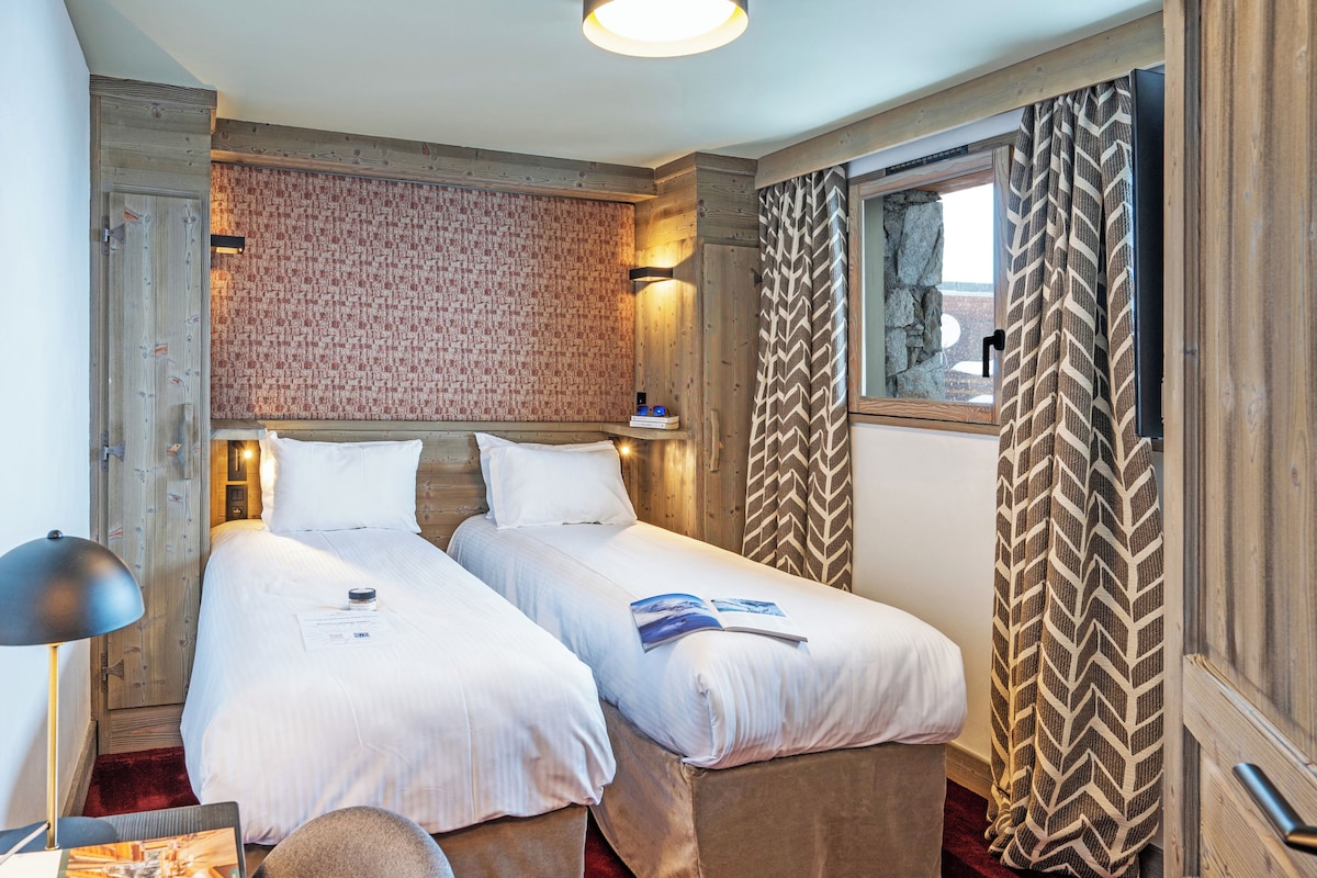 Habitación de hotel con dos camas, cabecero de madera, pared con diseño, ventana con cortinas y una lámpara.