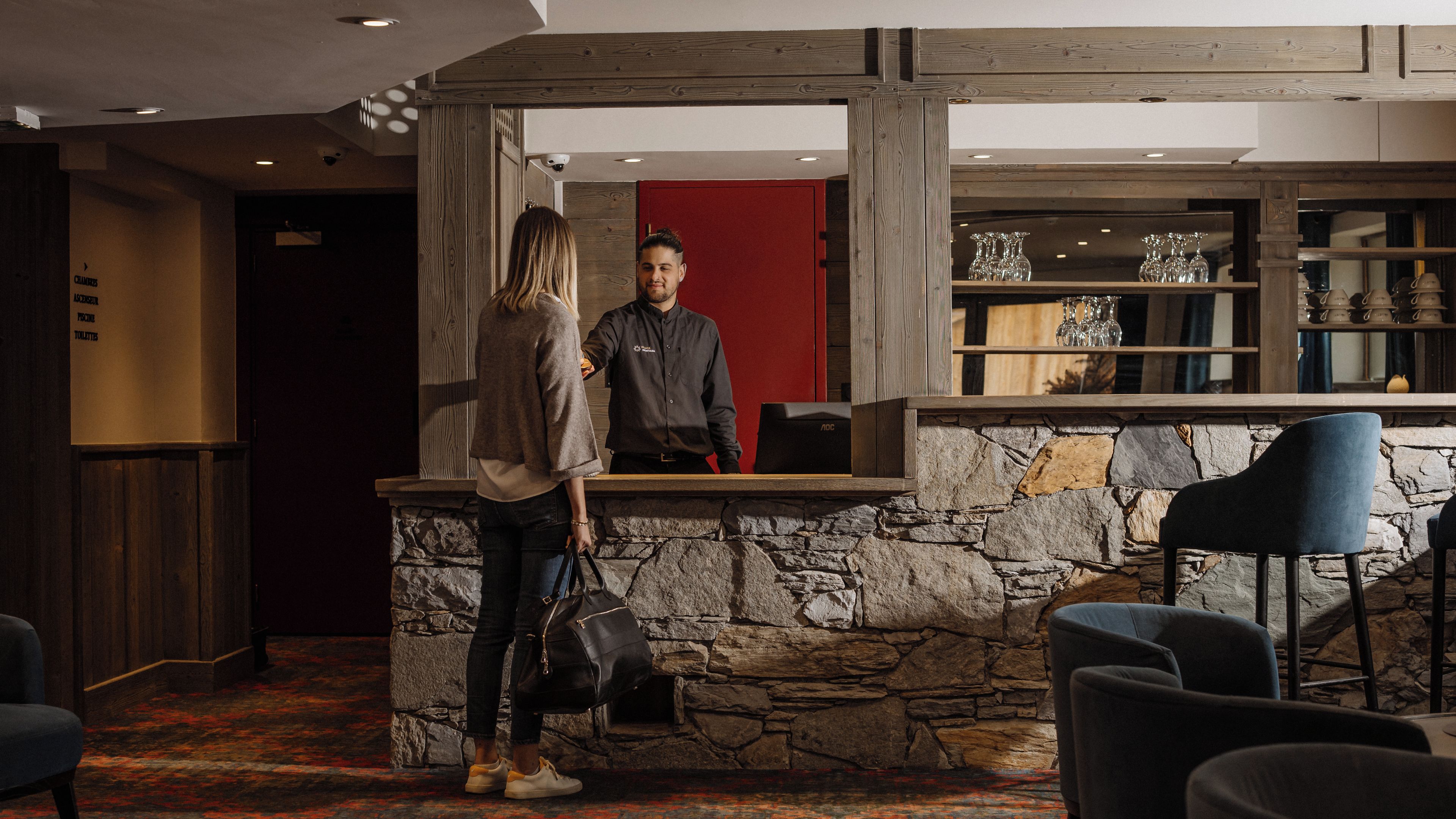 Un espace d’accueil avec un mur en pierre, une porte rouge et un comptoir de bar avec des chaises.