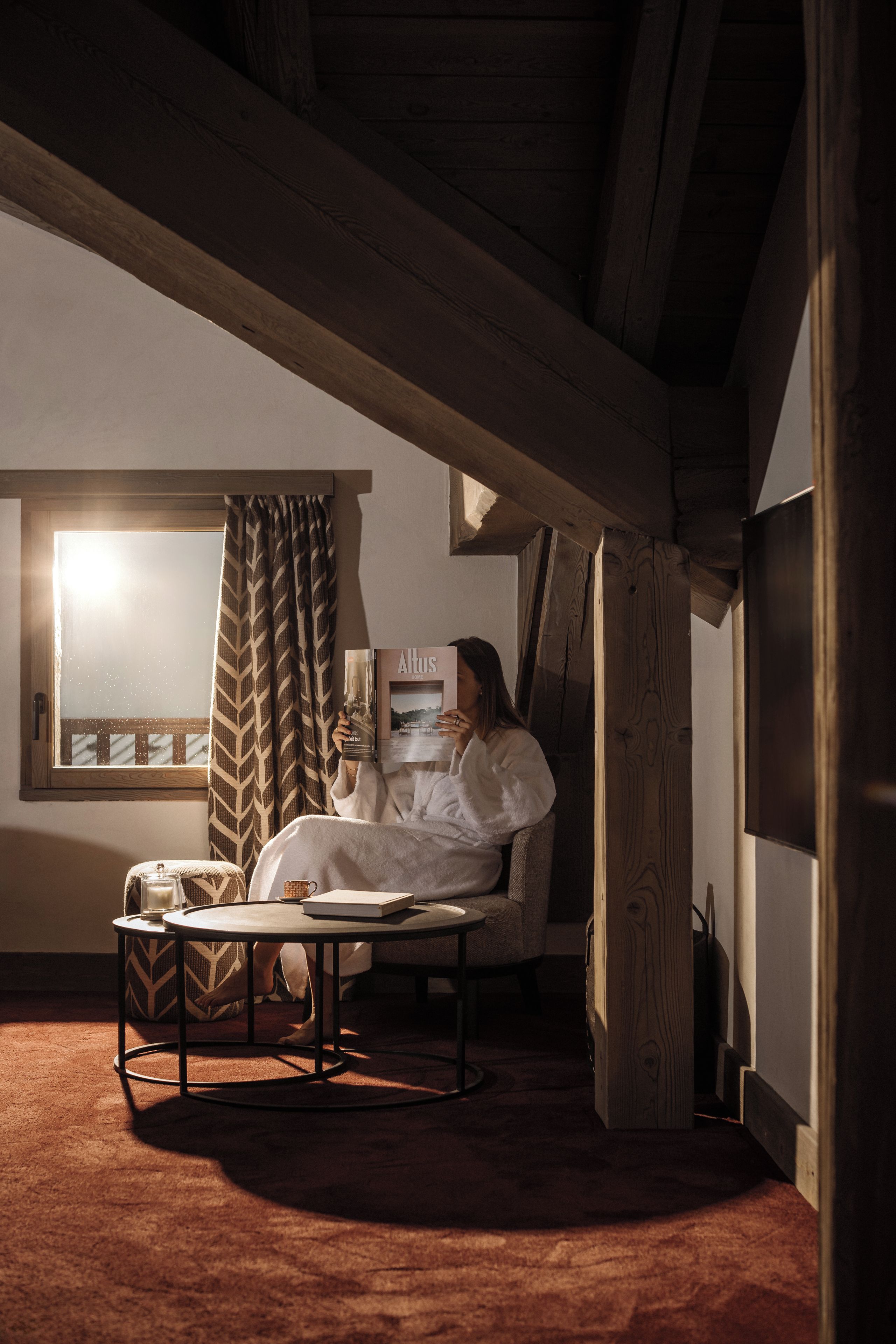 A cozy room with a wooden ceiling, a window with curtains, a chair, a table with a book, and a wall-mounted picture frame.