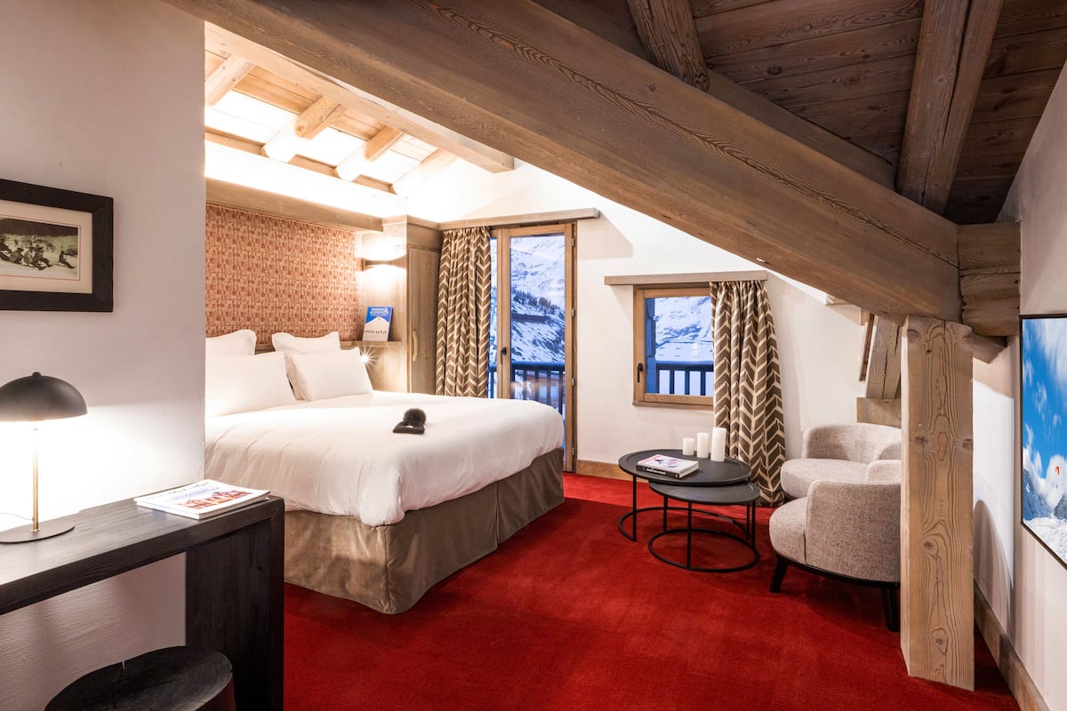 Habitación de hotel con una cama grande, un escritorio con lámpara, una silla y vistas a las montañas a través de la ventana.