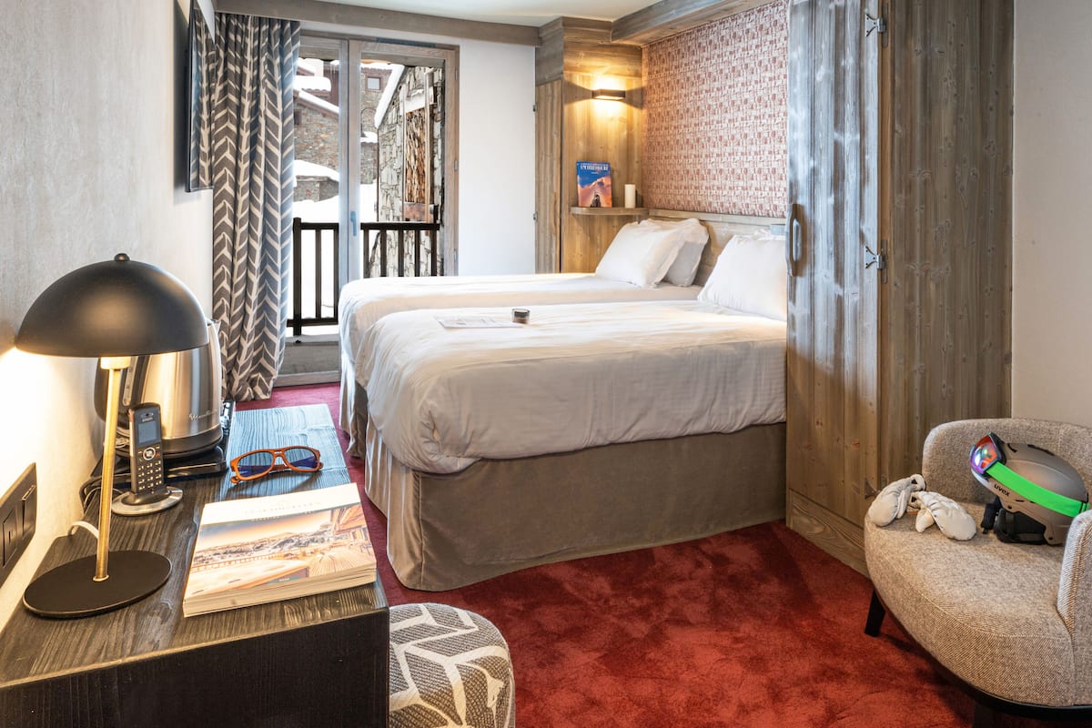 Habitación de hotel con dos camas, un escritorio con lámpara, una silla y una mesita con un libro y vasos.