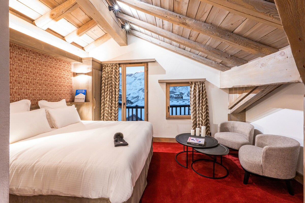 Habitación de hotel con una cama grande, una alfombra roja, una mesita con velas y vistas a una montaña nevada.