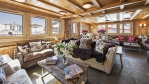 L’intérieur chaleureux d’un chalet, avec ses murs et son plafond en bois, de grandes fenêtres offrant une vue sur le paysage enneigé, et un coin salon confortable avec une table basse et des livres.