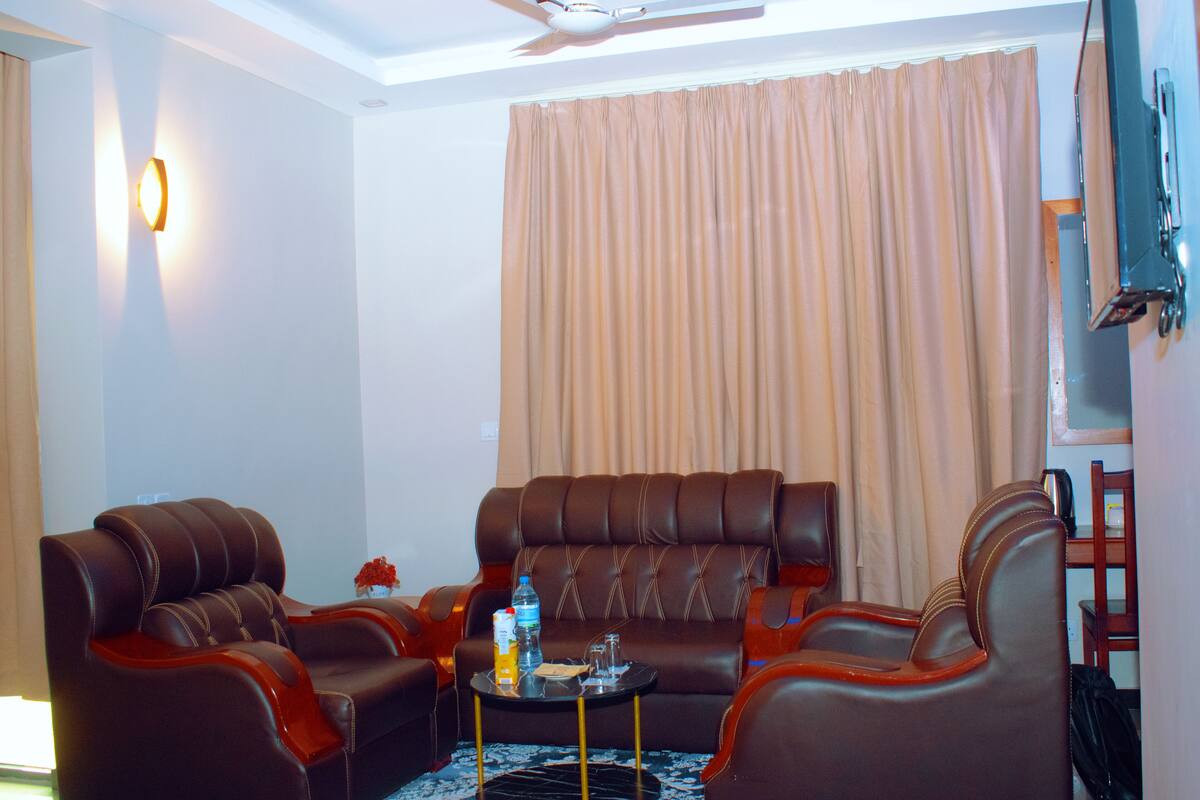 Une chambre d’hôtel avec un canapé en cuir marron, deux fauteuils, une petite table basse, un ventilateur de plafond, un téléviseur fixé au mur et une grande fenêtre avec des rideaux.
